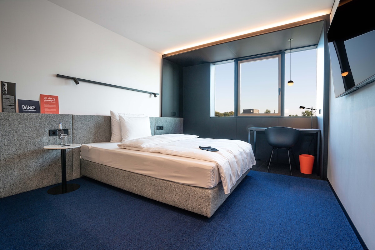 Ett modernt hotellrum med en säng, ett litet bord, en stol och ett fönster med utsikt.