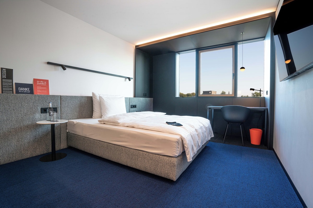 Ett modernt hotellrum med en säng, ett litet bord, en stol och ett fönster med utsikt.