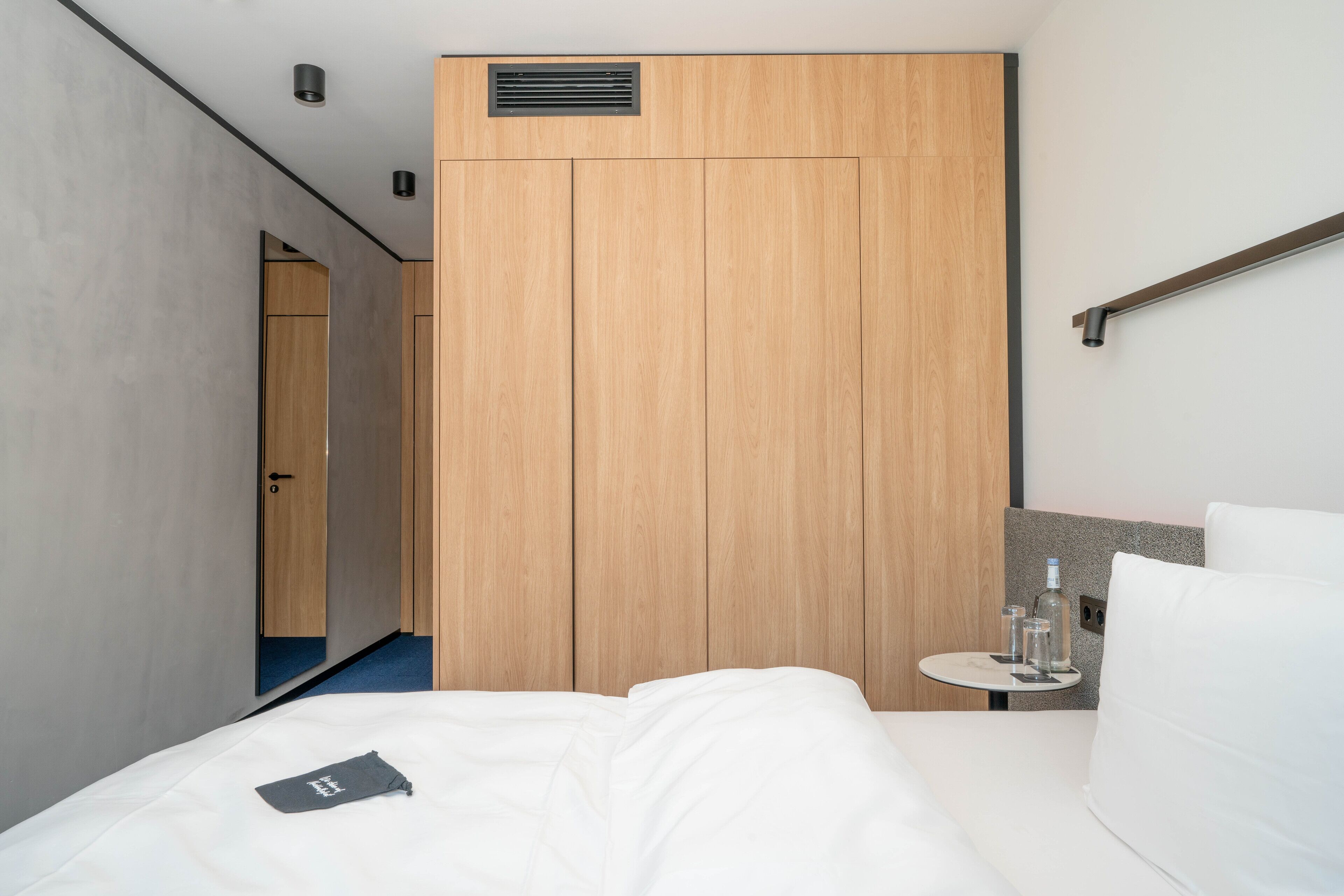 A modern hotel room with a large bed, a small bedside table with glasses, and a wooden wardrobe.
