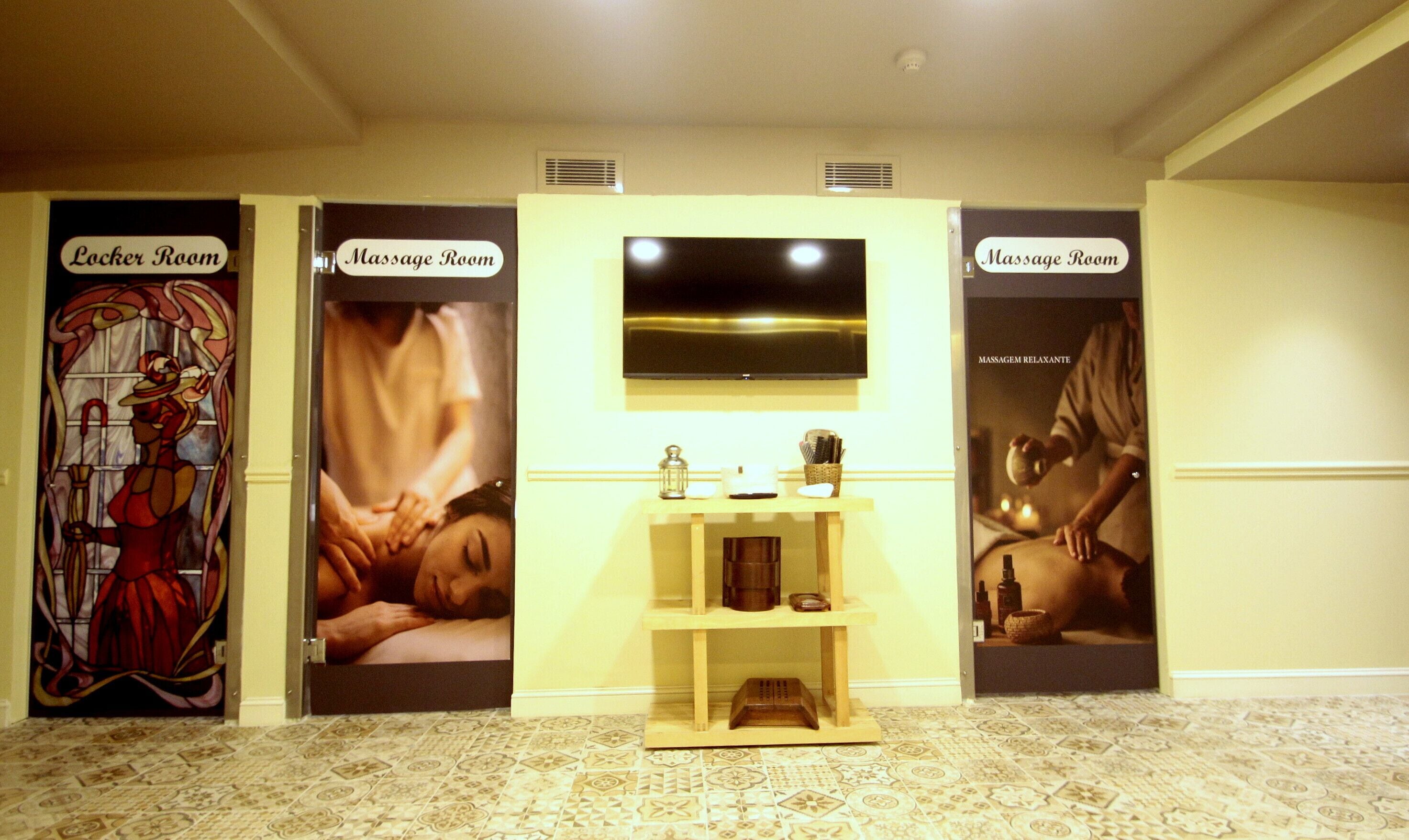 Un hall d’hôtel divisé en trois espaces distincts, identifiés par les panneaux « Salle des casiers », « Salle de massage  » et une autre « Salle de massage ». Chaque espace est doté d’un panneau indicateur et d’une grande affiche illustrant la fonction correspondante. Le hall est carrelé et possède une grande étagère en bois au centre, sur laquelle sont disposés divers objets.