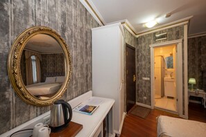 Habitación de hotel con un espejo grande y ornamentado, una mesa consola blanca con hervidor, cama con cabecera y baño visible a través de una puerta abierta.