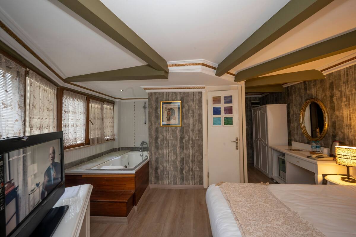 A hotel room with a large bed, a sitting area with a television, a bathroom with a bathtub, and a wooden floor.