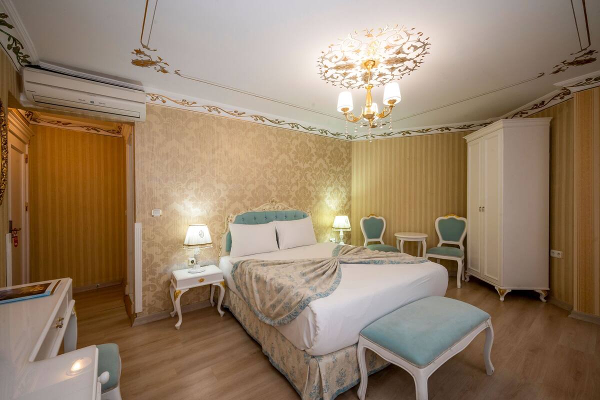 A hotel room with a large bed, two bedside tables with lamps, a chair, a wardrobe, and a chandelier.