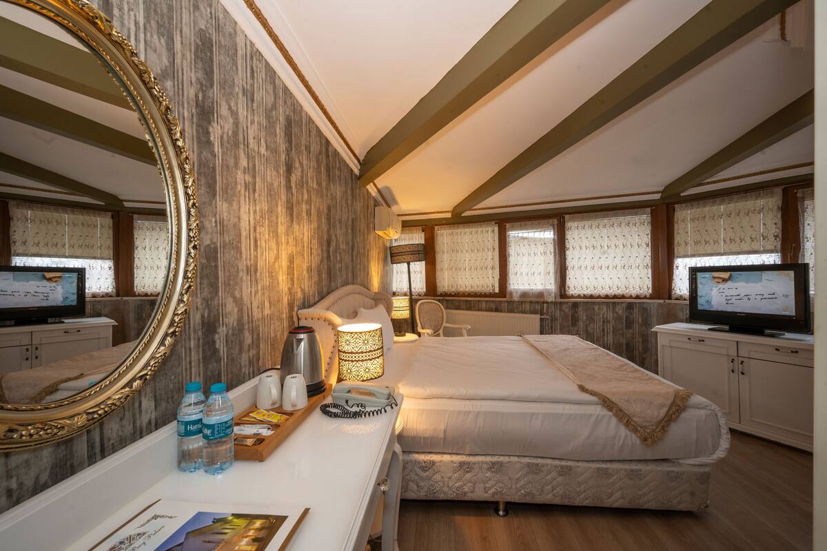 A hotel room with a large ornate mirror, a bed with a patterned duvet, a bedside table with a lamp, and a television.
