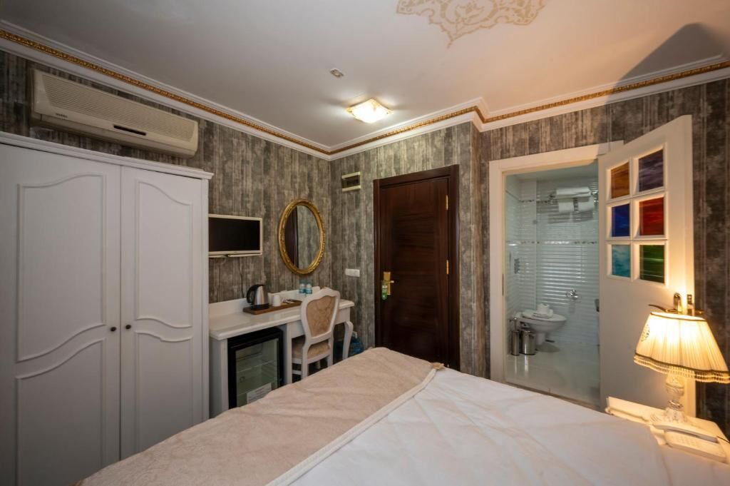 A hotel room with a bed, a desk, a chair, a wardrobe, a mirror, a bathroom with a toilet, and a lamp.