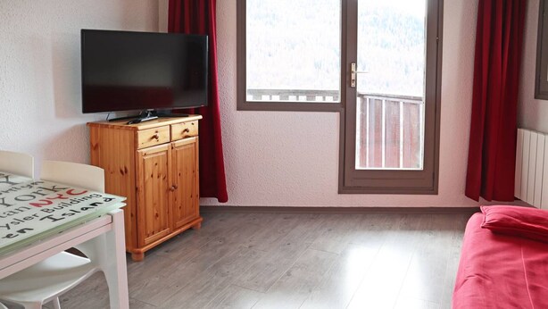 Une pièce avec un meuble en bois, un téléviseur, une table avec un set de table, un canapé rouge et une fenêtre avec des rideaux rouges.