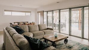 A modern living room with a sectional sofa, a wooden coffee table, and a view through large sliding glass doors to an outdoor patio area. - A Brand New Home with a Pool overlooking The Black Bull Golf Course (Yarrawonga)