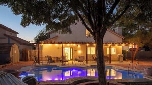 A backyard with a swimming pool, a covered patio, and a tree.