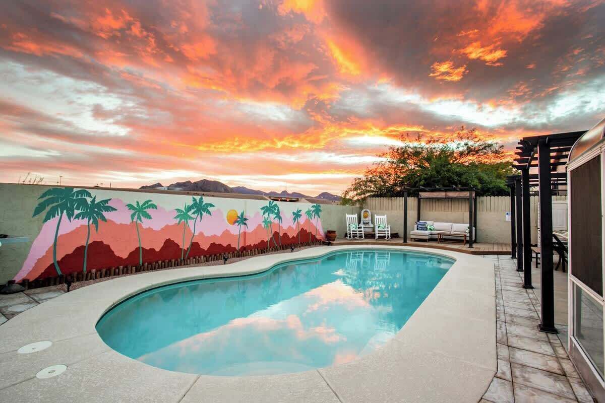 A swimming pool with a sunset mural, outdoor seating, and a view of mountains.