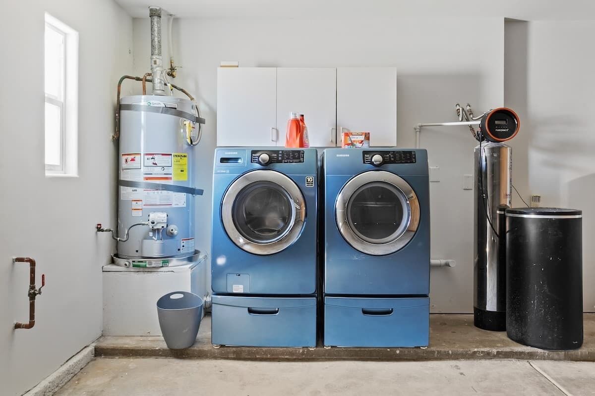 A laundry room with two blue front-loading washing machines, a water heater, and a trash can.