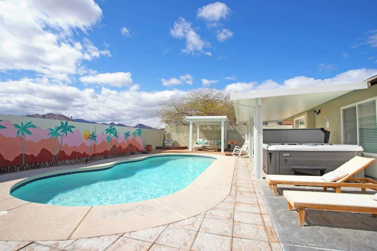 En swimmingpool med liggestole og jacuzzi, omgivet af en terrasse og et muralmaleri med palmer.