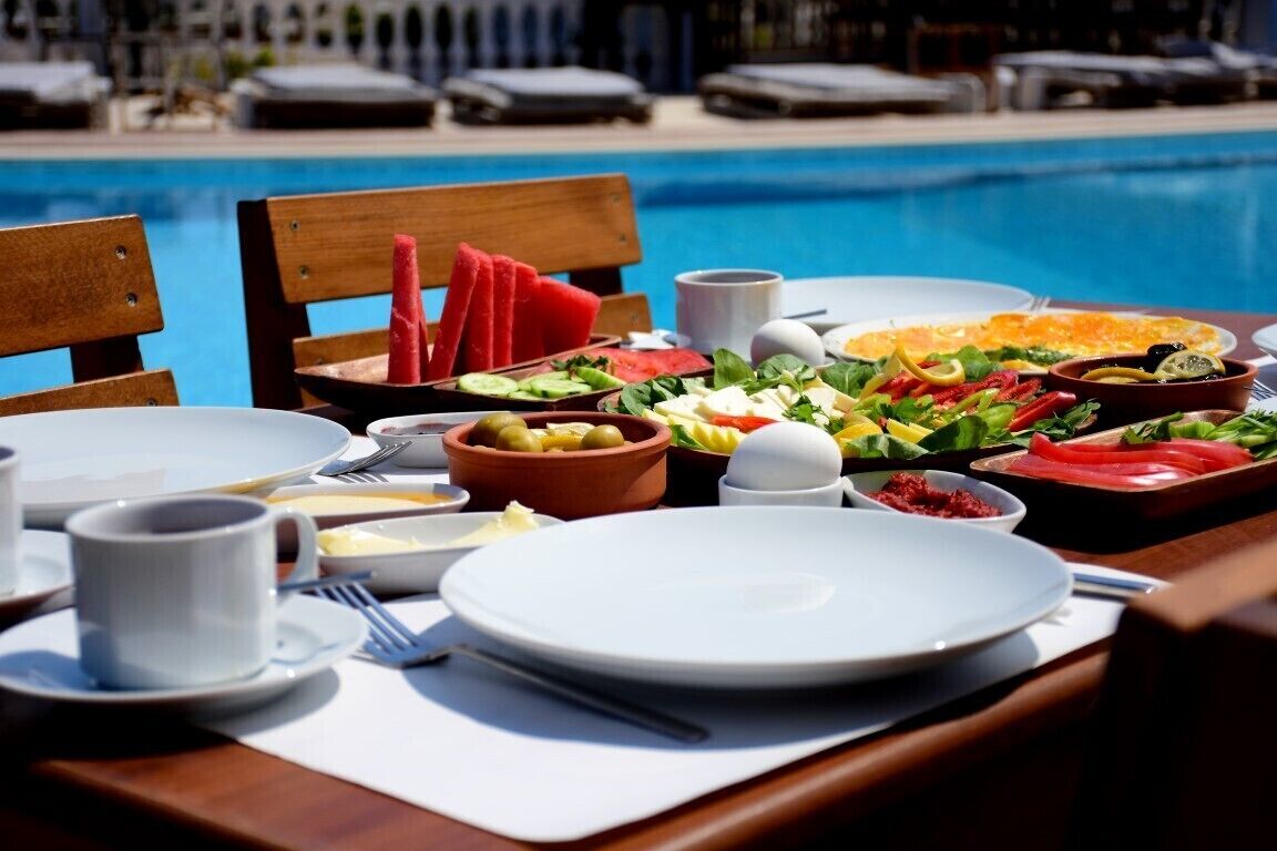 A dining table set for breakfast with various dishes, a pool in the background, and a wooden chair.