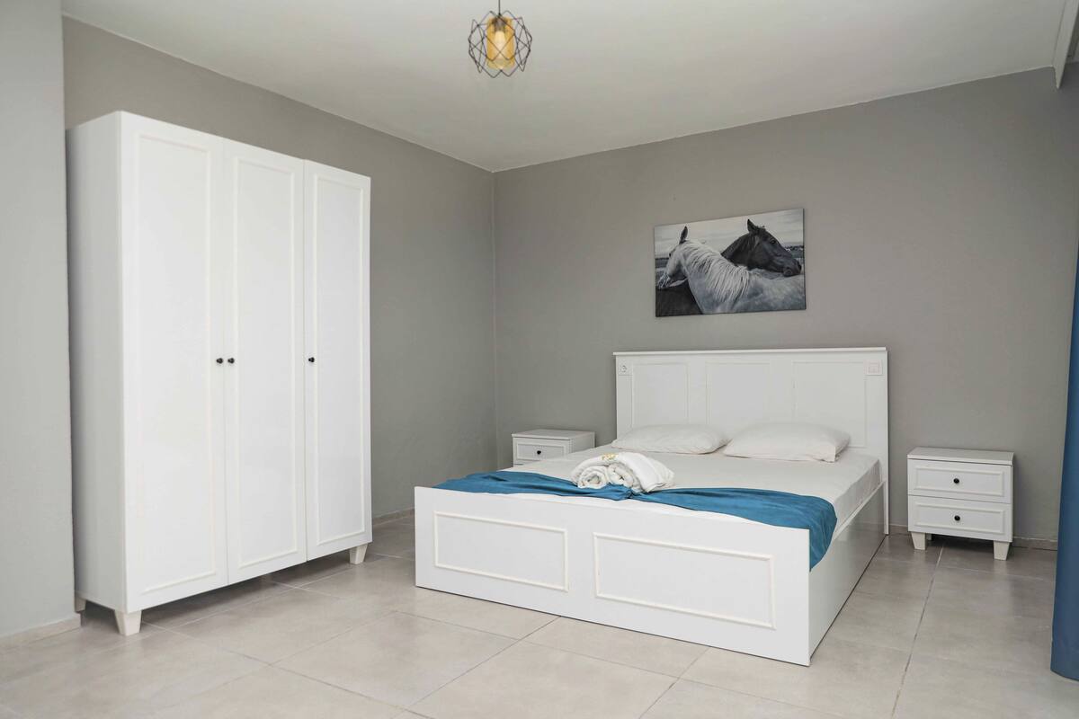 A bedroom with a large white wardrobe, a bed with a blue blanket, two bedside tables, and a wall-mounted picture of a horse.