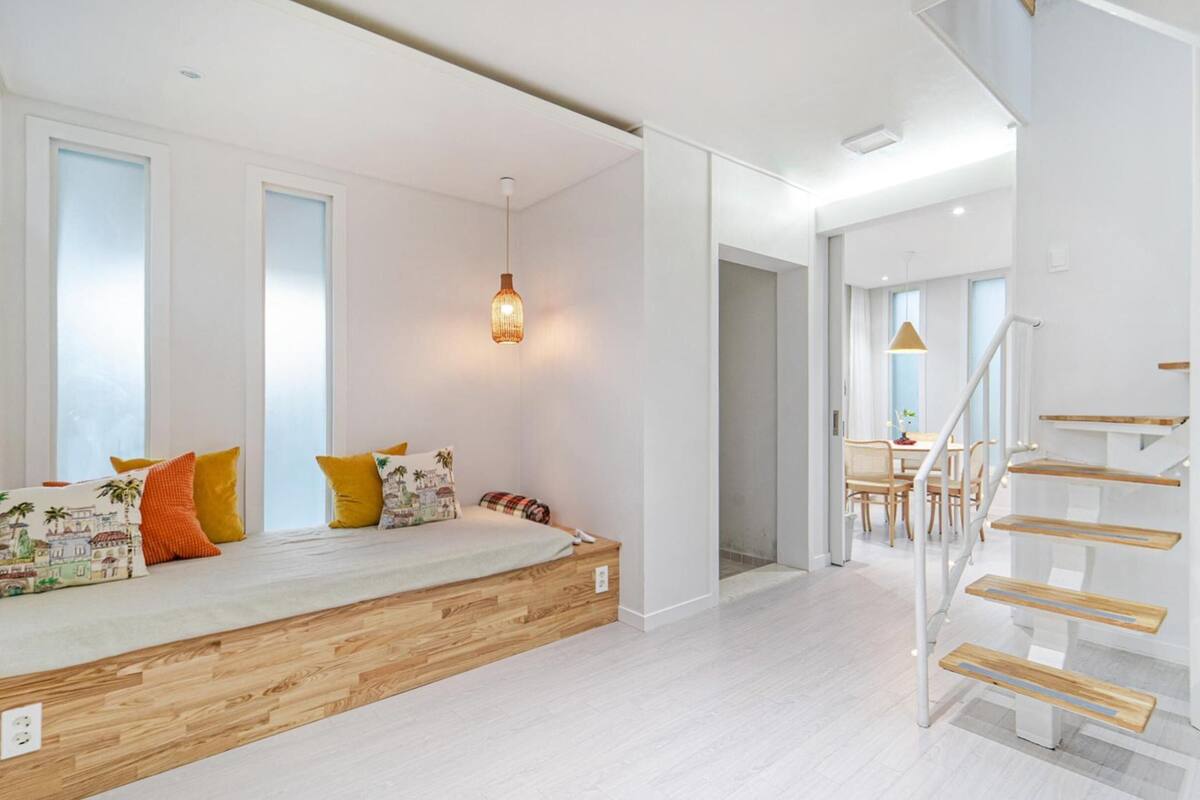 Ein heller, moderner Raum mit einem Bett aus Holzbank, einer Treppe mit Holzstufen und einem Essbereich im Hintergrund.