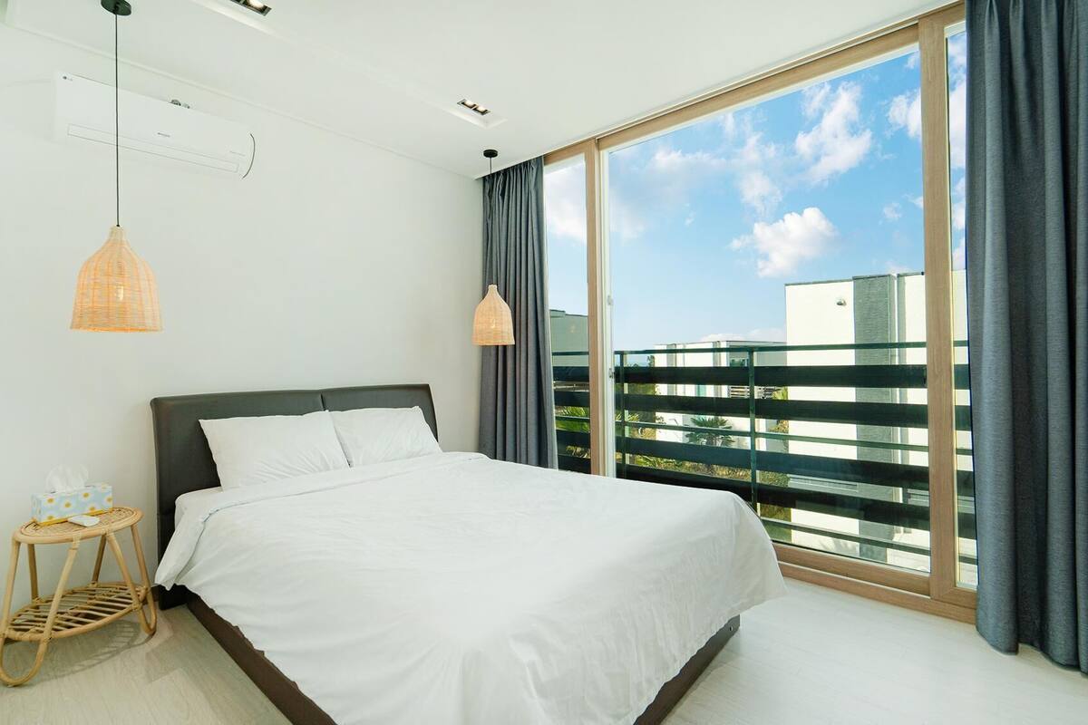 Ein modernes Schlafzimmer mit einem großen Bett, einem kleinen Nachttisch, einem Korbstuhl und einem Balkon mit Stadtblick.