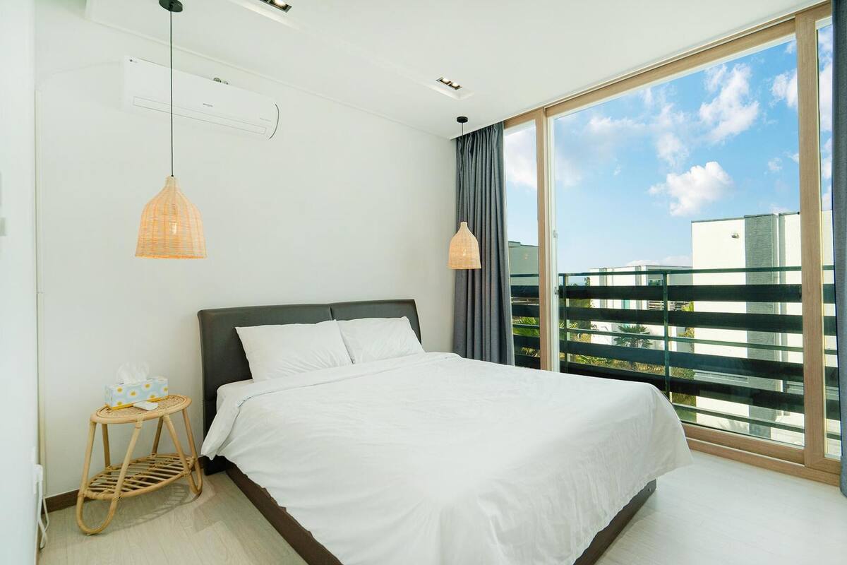 Ein modernes Schlafzimmer mit einem großen Bett, einem kleinen Nachttisch und Blick auf einen Balkon mit Geländer.