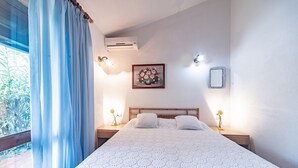 A bedroom with a large bed, two bedside tables with lamps, a wall-mounted air conditioner, a framed floral artwork, and a window with curtains.