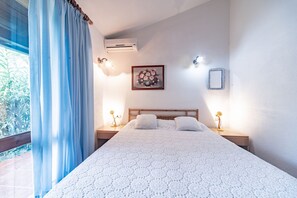 A bedroom with a large bed, two bedside tables with lamps, a wall-mounted air conditioner, a framed floral artwork, and a window with curtains.
