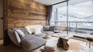 Ein modernes Wohnzimmer mit einer Holzwand, einem grauen Ecksofa, einem runden Couchtisch und Blick auf verschneite Berge durch große Fenster.