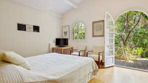Une chambre avec un lit, une télévision, un bureau et des chaises. Il y a une grande fenêtre et une porte ouverte qui donne sur un jardin.
