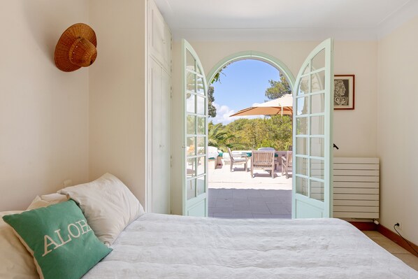 A bedroom with a bed, a pillow with "ALOHA" on it, and an open door leading to a patio with a chair and umbrella.