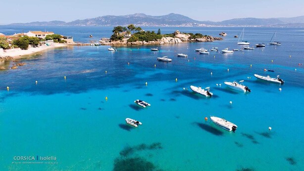 Une zone côtière où des bateaux sont amarrés dans une eau d’un bleu limpide, une côte rocheuse bordée de bâtiments et, au loin, des montagnes.