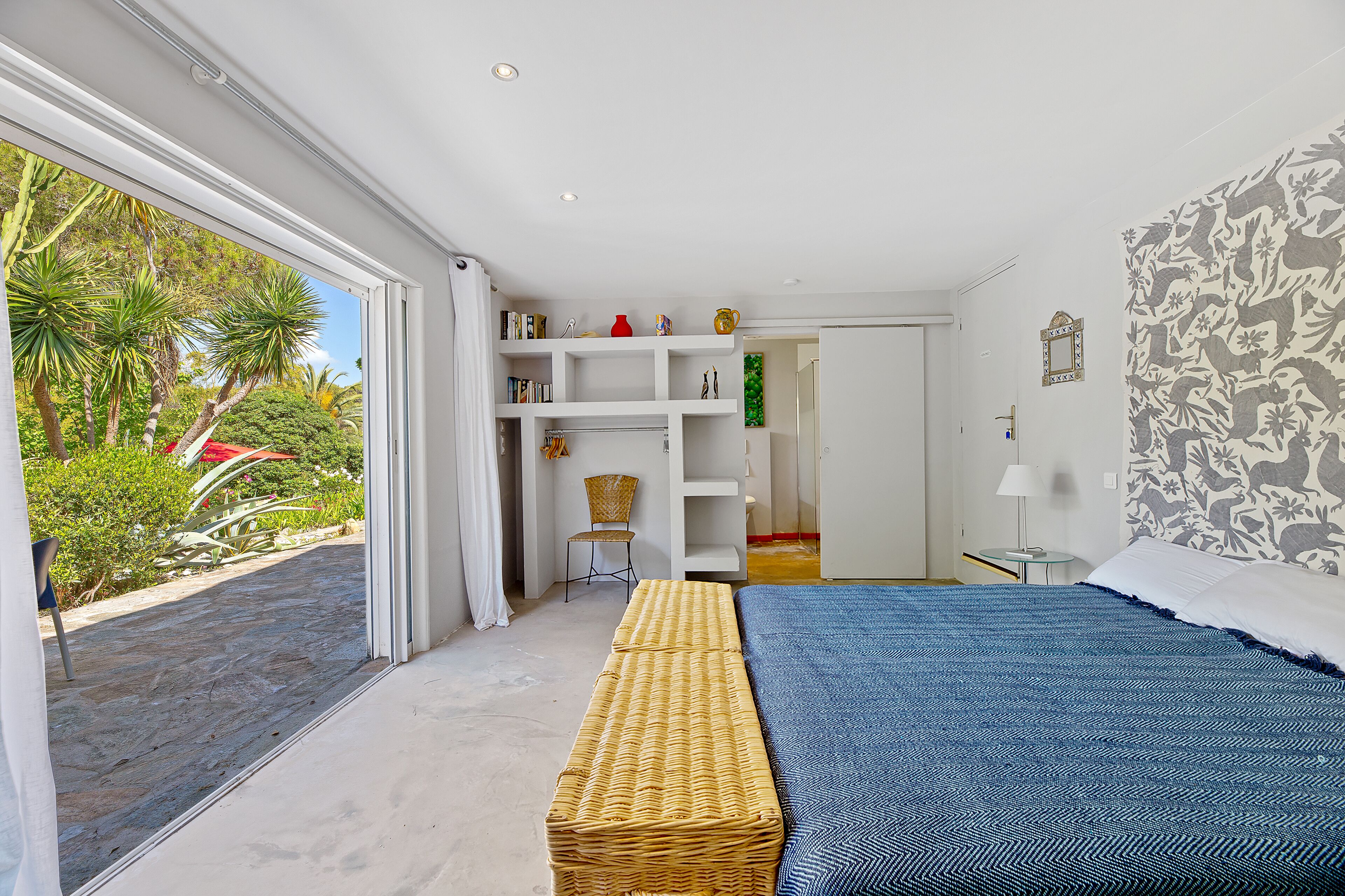 A bedroom with a large bed, a bench, a white wardrobe, and a view of the outdoors.