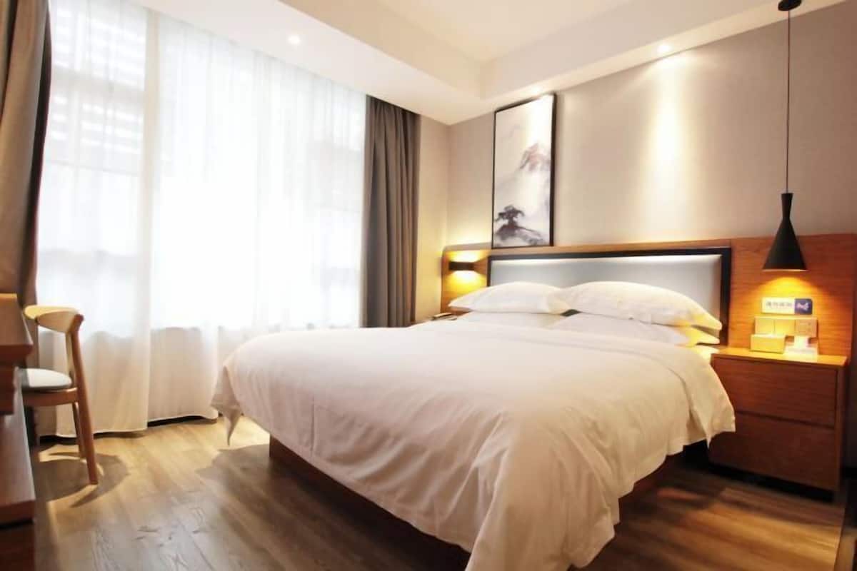 A hotel room with a large bed, a wooden nightstand with a lamp, a chair, and a window with curtains.