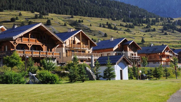 A row of wooden chalets with sloped roofs, set against a backdrop of green hills and mountains. - Studio for 4 with terrace, garden and parking (Montgenèvre)