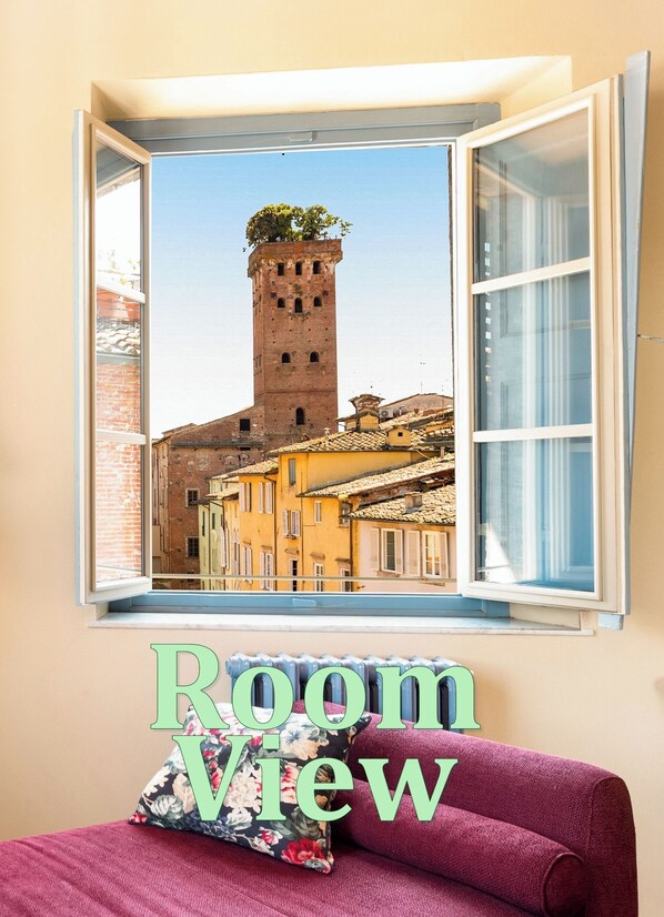 Ein Zimmer mit Fensterblick auf einen historischen Turm und Gebäude, einem roten Sofa und einem floralen Kissen.