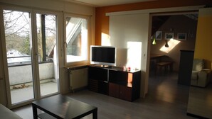 Sala de estar con televisor, sofá y una mesa. Hay un ventanal corredero que da a un balcón.