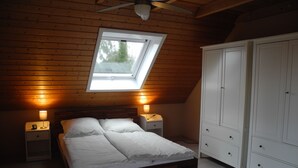 Un dormitorio con techo de madera, tragaluz, cama con ropa de cama blanca, dos mesitas de noche con lámparas y un armario.