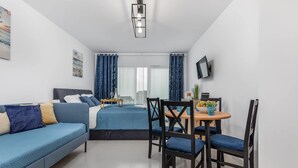 A modern room with a bed, a sofa, a dining table with chairs, a TV mounted on the wall, and a window with curtains.