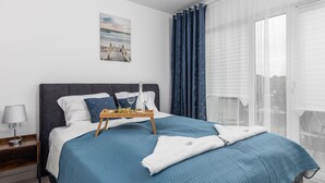 A neatly made bed with a blue bedspread, a bedside table with a lamp, and a window with sheer curtains.