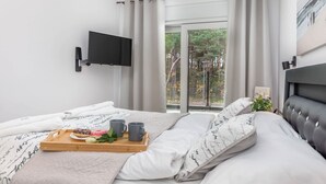 Una camera da letto moderna con un letto, una TV fissata al muro, un balcone con vista sugli alberi e un vassoio con ciambelle e tazze da caffè.
