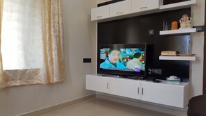 A living room with a flat-screen TV on a white entertainment unit, a window with sheer curtains, and a shelf with decorative items.