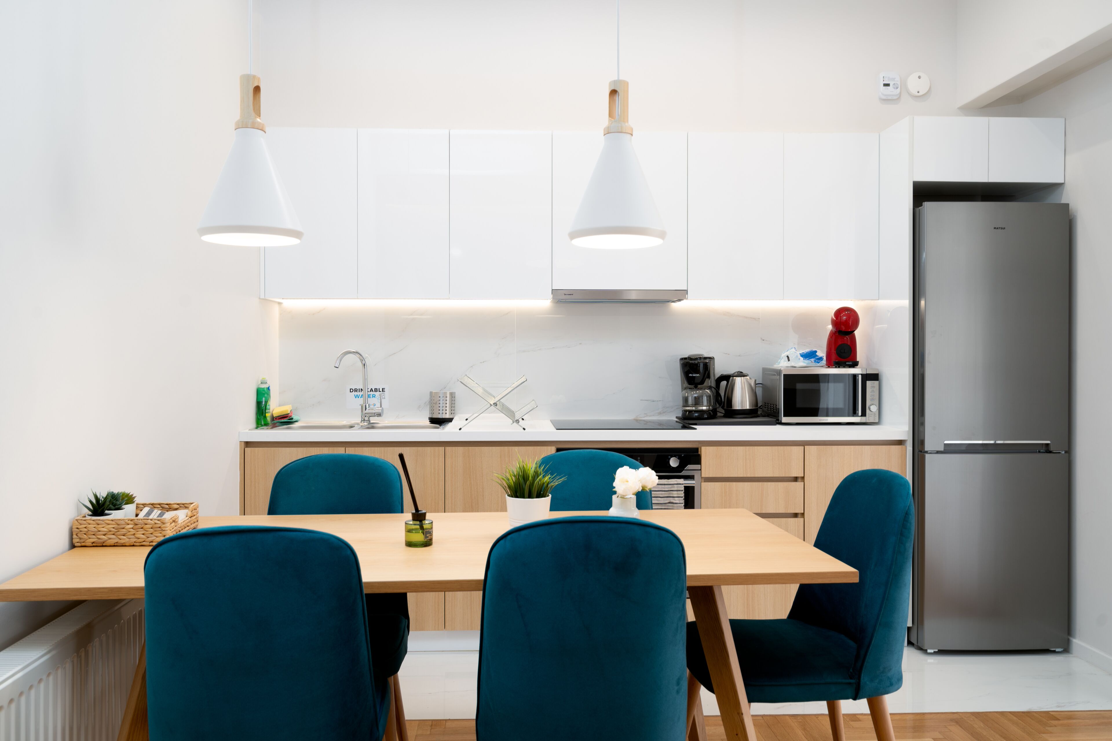 A modern kitchen with white cabinets, a stainless steel refrigerator, and teal chairs around a wooden table.