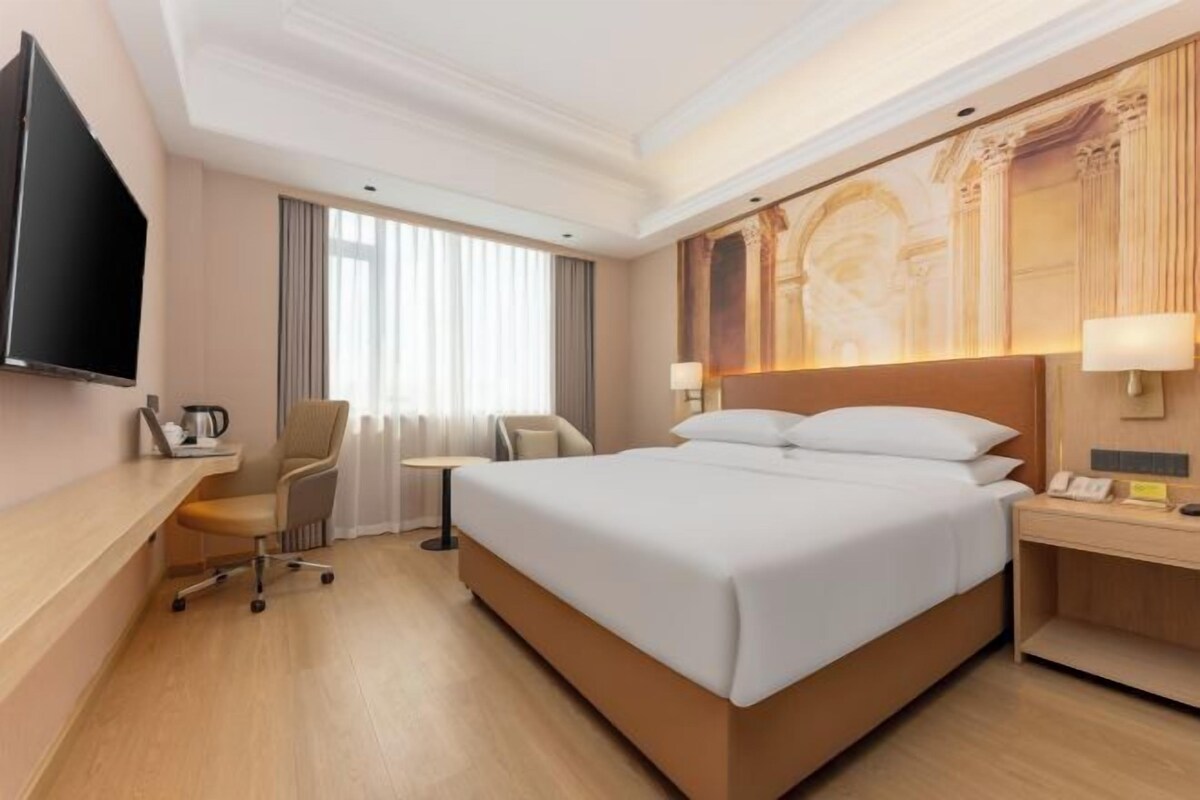A hotel room with a large bed, a desk, a chair, a television, and a window with curtains.