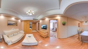 Un salón espacioso con un sofá grande, una mesa de centro y un televisor. La habitación tiene piso de madera y un techo alto con molduras decorativas.