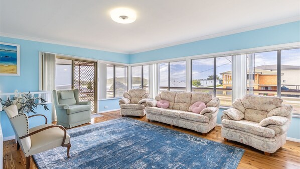 Relax in this spacious living room with a large window offering a view of the surrounding area, a light-colored sofa set, and a single armchair for comfortable seating.