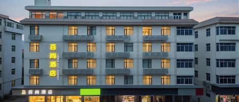 A multi-story hotel with a row of balconies on each floor, a ground-level entrance with a green sign, and a clear sky in the background.