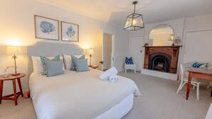 A bedroom with a large bed, two bedside tables with lamps, a chair, a fireplace, and framed artwork on the wall.
