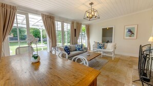 A spacious living room with a wooden table, white wicker sofa, and a view of the garden through large windows.