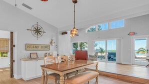Een ruime eethoek met een houten tafel en bank, witte kasten en een groot raam met uitzicht op het strand.