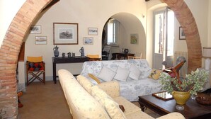A living room with a sofa, armchairs, a coffee table, and a dining table. There are framed pictures on the wall and a potted plant on the table.
