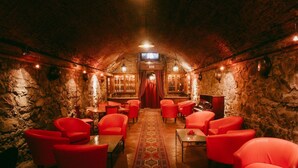 Ein gemütlicher, schummrig beleuchteter Raum mit Steinwänden, rot gepolsterten Sesseln, einem gemusterten Teppich und einer Bar mit Flaschen.