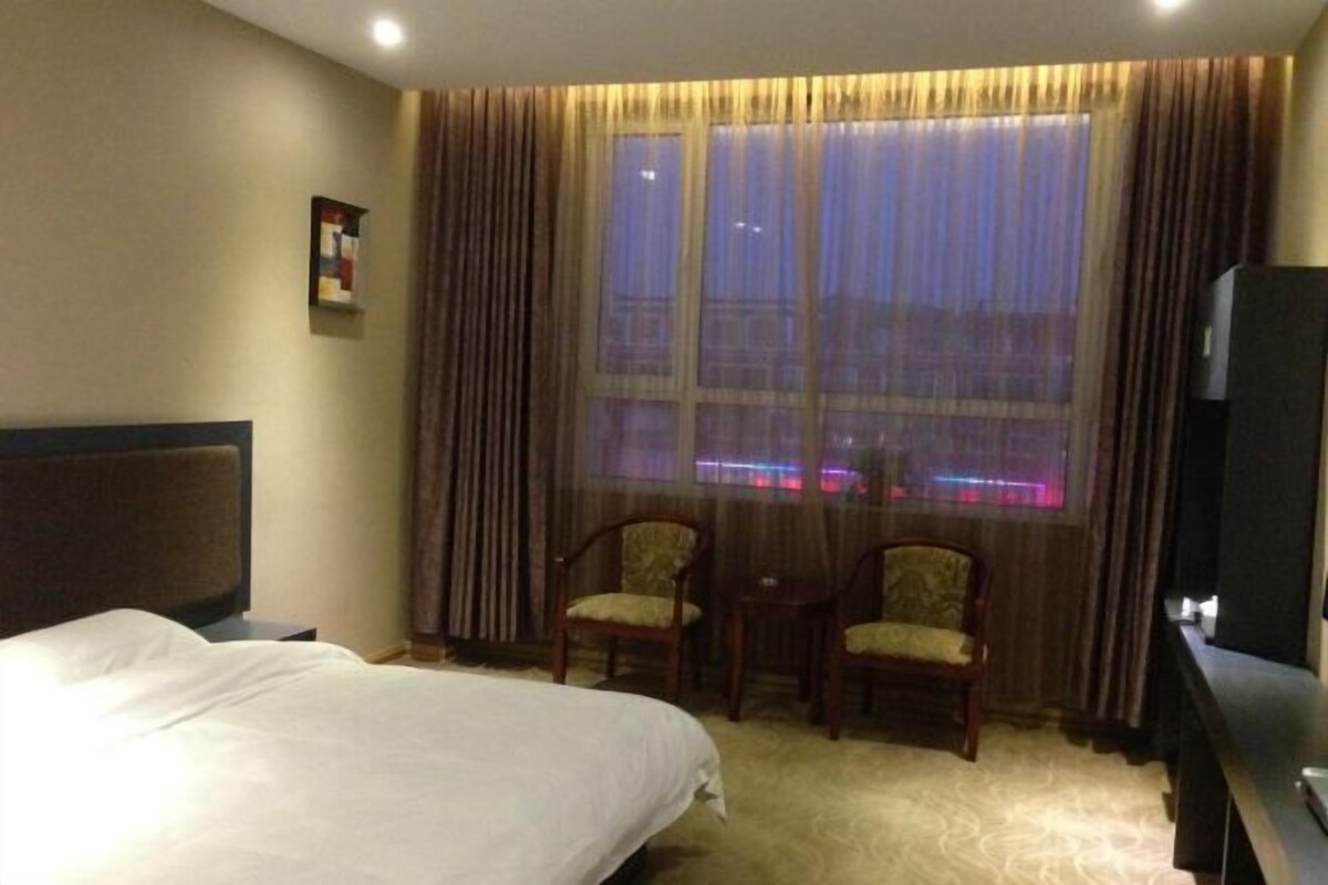 Ein Hotelzimmer mit einem Bett, zwei Stühlen, einem kleinen Tisch, einem Fernseher und einem Fenster mit Vorhängen.