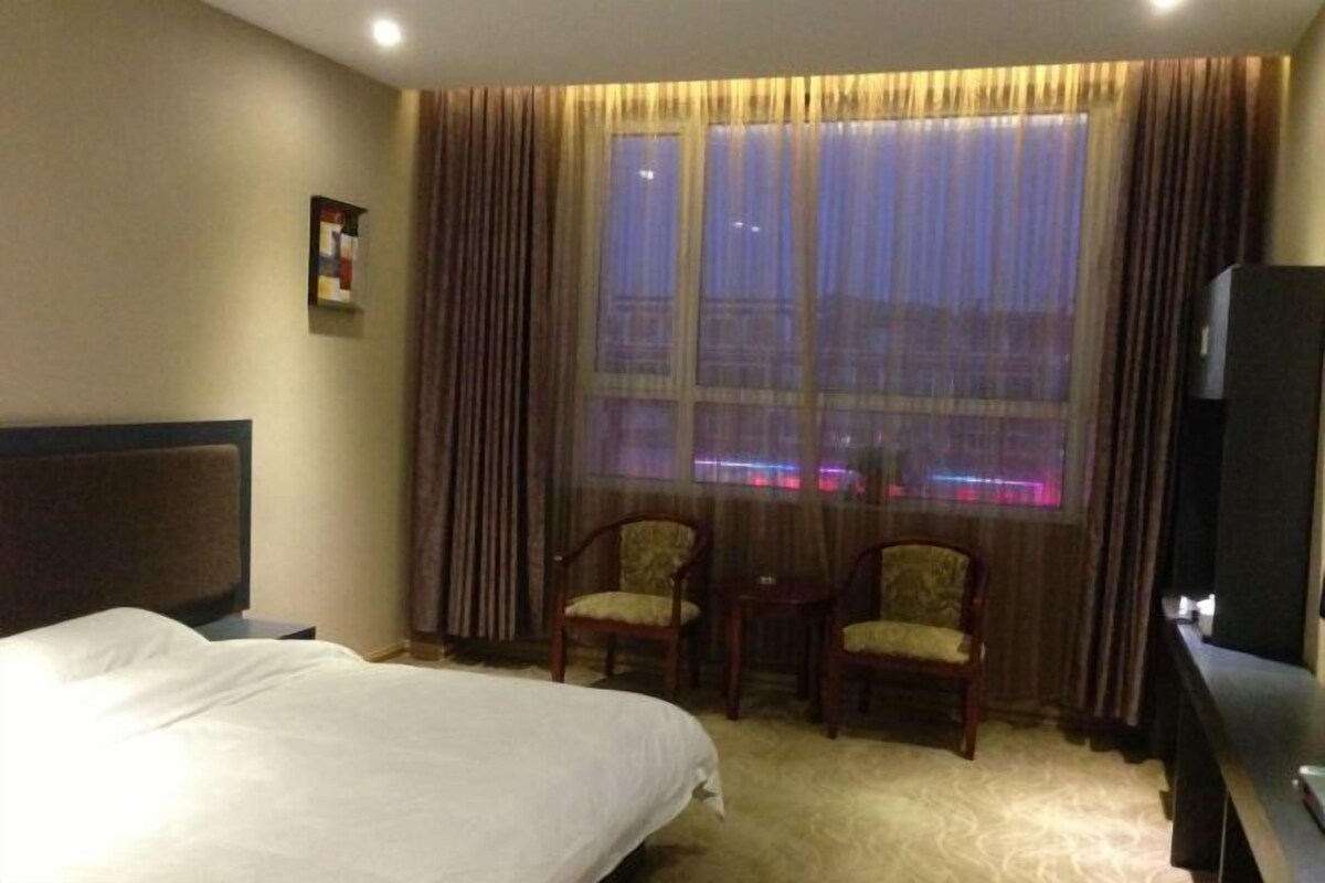 Ein Hotelzimmer mit einem Bett, zwei Stühlen, einem kleinen Tisch, einem Fernseher und einem Fenster mit Vorhängen.