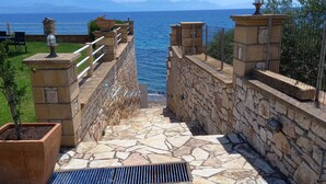 Uma escadaria de pedra leva a uma varanda com vista para o mar, com grades de metal e plantas em vasos.