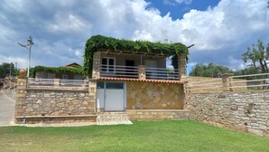 Uma casa de dois andares com fachada de pedra, uma varanda com grades de metal e um pequeno jardim frontal.